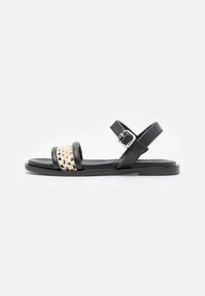 Sort flad sandal med en beige vævet frontrem, en justerbar ankelstrop med en sølvspænde og en tynd sål.