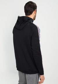 Svart huvtröja av mjukt tyg med huva, vita och färgade mönster längs ärmarna samt ribbade muddar i midjan.