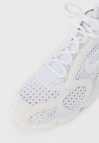 Gros plan d'une chaussure de sport blanche avec tige en mesh, lacets blancs et logo Swoosh sur le côté, sur fond blanc.