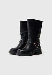 Par de botas de piel negras hasta la mitad de la pantorrilla con tacones cuadrados y correas decorativas que presentan anillos y tachuelas de metal dorado.