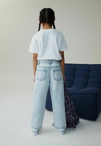 Tricou alb, blugi largi de culoare albastru deschis și adidași. Persoana ține o cămașă în carouri, văzută din spate, într-un cadru neutru.