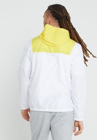White lightweight jacket with a yellow hood and back yoke. Elastic cuffs and hem, featuring a smooth texture and modern design.