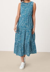 Une robe sans manches couleur sarcelle avec un motif de feuilles, une jupe à volants et un tissu texturé, associée à des baskets blanches.