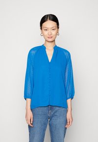 Blusa azul com mangas semi-transparentes de três quartos, textura de bolinhas, decote em V e fecho frontal por botões. Combinada com jeans azuis.