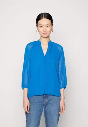 Blusa azul com mangas semi-transparentes de três quartos, textura de bolinhas, decote em V e fecho frontal por botões. Combinada com jeans azuis.