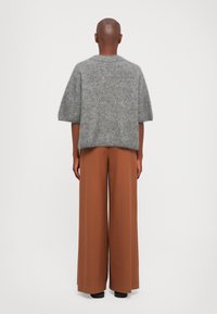 Personne avec la tête rasée portant un pull gris en peluche à manches courtes et un large pantalon marron, se tenant devant un fond blanc uni.