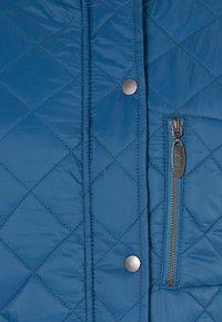 Quiltet blå jakke med en glänsende finish, sølvfarvede trykknapper og en sidezipper med en struktureret trækstang til lukning.