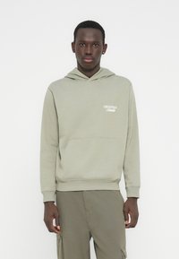 Νεαρός άνδρας με ανοιχτό πράσινο φούτερ με κουκούλα που έχει το κείμενο «Originals Studio» και ταιριαστό παντελόνι cargo, στέκεται μπροστά σε λευκό φόντο.