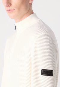 White ribbed pullover with a short zip collar. Features a black fabric patch with "Barbour International" on the left sleeve. Textured design.