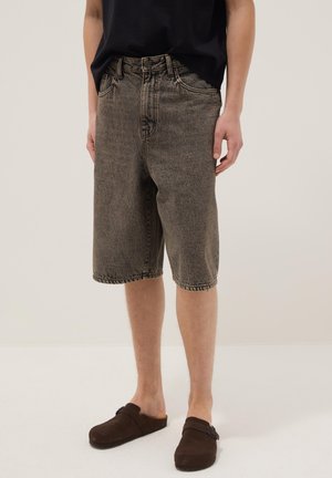 Persona con pantaloncini in denim scuro lavato fino al ginocchio, maglietta nera e scarpe marroni slip-on con fibbia, in piedi su un pavimento chiaro.