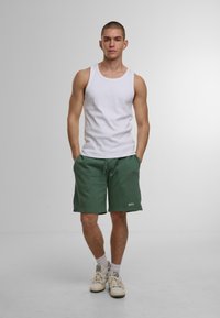 Débardeur blanc sans manches et short vert avec cordon de serrage. Le short a un petit détail textuel. Le mannequin porte des baskets blanches avec des accents gris.