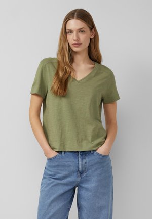 Frau mit langen blonden Haaren, die ein grünes kurzärmeliges V-Neck-Shirt und hellblaue Jeans trägt, steht mit den Händen in den Taschen.