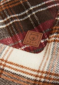 Tissu à carreaux aux tons marron, rouge, crème et orange avec une pièce en cuir cousue représentant un visage d'ours et le mot NEXT.