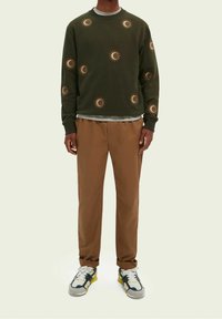 Olivengrüner Pullover mit Sonnenmotiven, kombiniert mit braunen Hosen und mehrfarbigen Turnschuhen. Strukturierter Stoff, Rundhalsausschnitt.