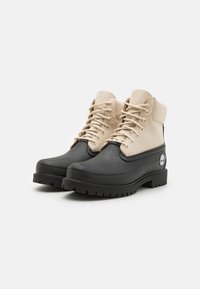 Botas de tobillo de goma negra y cuero crema con cordones, parte superior de cuero texturizado, puntera redonda y suela de goma negra estriada. Detalle del logo de Timberland.