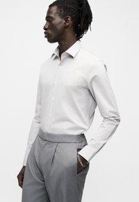 Άνδρας με κοτσιδάκια, φοράει λευκό πουκάμισο με κουμπιά και γκρι πλισέ παντελόνια, ποζάρει με το ένα χέρι στην τσέπη μπροστά σε λευκό φόντο.