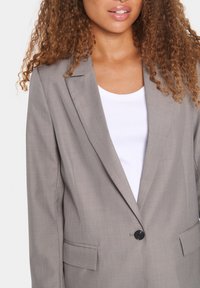 Šedý blazer s hladkou texturou a jedním černým knoflíkem. Má výstřih do V a boční kapsy, nošený přes bílý top.