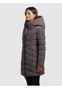 Abrigo acolchado largo y gris con una gran capucha, cierre con cremallera en la parte delantera y patrón de costura en chevrón. Presenta una marca negra y bolsillos laterales.
