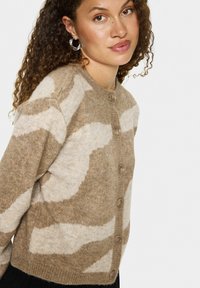 Bežs vilnas kardigans ar gaiši brūnu abstraktu rakstu, apaļo apkakli, priekšējiem pogām un ribotu apakšmalu, kas redzams uz modeļa ar cirtainiem matiem.