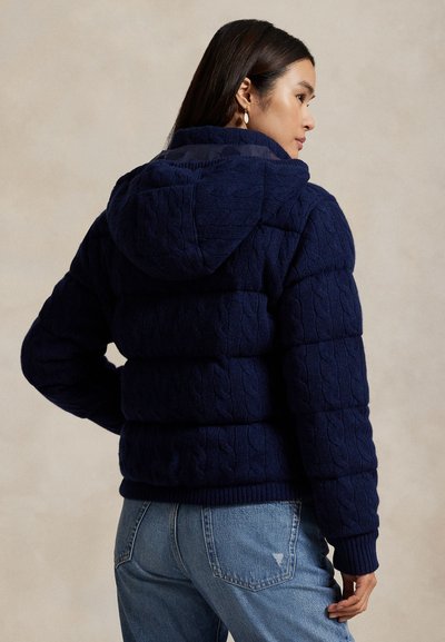 Marinblå kabelstickad puffjacka med huva, med texturerade paneler och ribbade ärmar. Bärs tillsammans med ljusblå jeans.