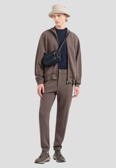 Veste zippée marron, t-shirt bleu marine, pantalon marron assorti et baskets grises. Un chapeau seau beige clair et un petit sac bandoulière noir complètent le look.