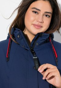 Giacca impermeabile blu navy con chiusura a zip, cordini rossi e cappuccio removibile, dotata di una etichetta con logo vicino alla zip.