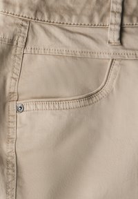 Beige látková kapsa na kalhotách s viditelným stehem a kovovými detaily. Hladká textura, čisté linie a dobře padnoucí design.