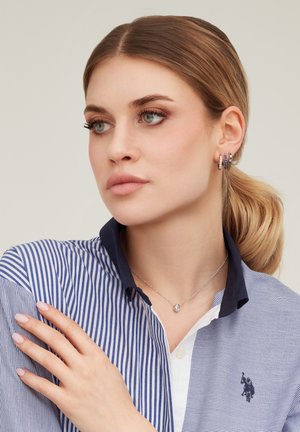 Giovane donna con capelli biondi raccolti in una coda bassa, che indossa una camicia a righe, una collana d’argento con pendente a goccia e diversi orecchini, guarda di lato.