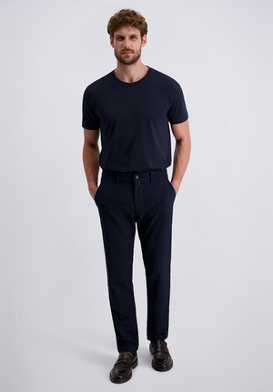 Mann mit lockigem Haar, der ein eng anliegendes dunkles marineblaues T-Shirt, passende Hose und dunkelbraune Leder-Schlüpfschuhe trägt und mit den Händen in den Taschen steht.