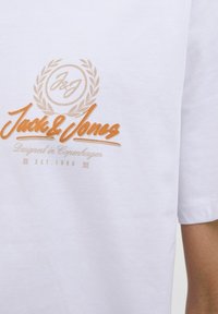 Balti kokvilnas T-krekls ar oranžu un brūnu logotipu, kurā iekļauts teksts "Jack & Jones", kā arī dekoratīvi elementi, piemēram, lauru lapas.