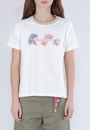 Žena v bílém tričku s květinovými vzory a nápisem „Cafe Noir“, v kombinaci se zelenými kalhotami a růžovou klíčenkou visící z kapsy.