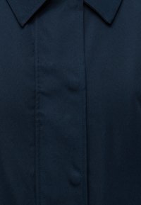 Gros plan d'un col de chemise bleu foncé avec une patte de boutonnage cachée et des coutures visibles sur le panneau avant.