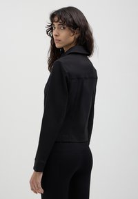 Donna con capelli ricci scuri guarda oltre la spalla indossando una giacca nera a maniche lunghe e pantaloni neri a coste su uno sfondo semplice.
