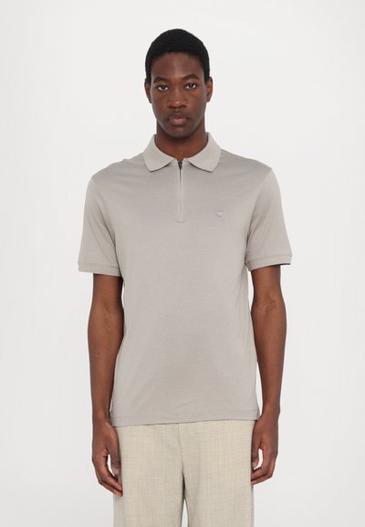 Polo shirt beige con zip al collo e maniche corte, realizzato in materiale morbido, con un logo discreto sul petto sinistro e una texture semplice.