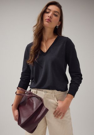 Hosszú barna hajú nő fekete V-nyakú pulóvert és bézs, csíkos nadrágot visel, kezében nagy, sötétlila bőr táska van az oldalán.