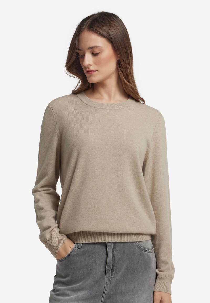 Beżowy dziany sweter z okrągłym dekoltem, długimi rękawami i luźnym krojem. Miękka faktura, ściągacze na mankietach i dole. Połączony z szarymi spodniami.