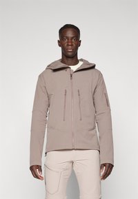 Svijetlosmeđa jakna od softshell materijala s kapuljačom, dvije prednje zip džepove i logotipom na rukavu. Ima glatku teksturu i prilagođeni kroj.