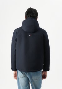 Navy blue hooded jacket with a smooth texture, featuring a subtle logo detail on the back and a relaxed fit, paired with light denim jeans.