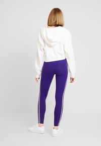 Vit kortad hoodie med huva, kombinerad med lila leggings med vita sidostripes. Modell bär vita träningsskor. Enfärgad bakgrund.