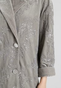 Chaqueta de terciopelo gris con bordados florales y lentejuelas. Presenta un gran cuello, dos botones y mangas con puños. Textura suave con acentos detallados.