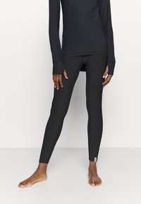 Leggings neri realizzati in materiale elasticizzato, design aderente, texture liscia e un piccolo etichetta di marca alla caviglia, indossati con un top a maniche lunghe.