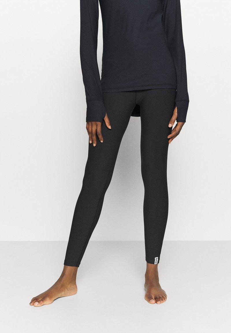 Leggings neri realizzati in materiale elasticizzato, design aderente, texture liscia e un piccolo etichetta di marca alla caviglia, indossati con un top a maniche lunghe.