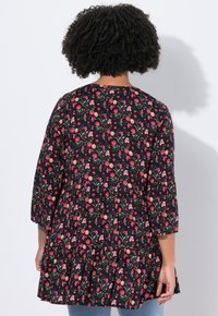 Femme avec des cheveux bouclés portant une robe fleurie noire avec des fleurs roses, rouges et oranges, se tenant contre un fond clair.