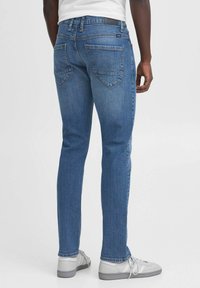 Jeans ajustados de color azul con cinco bolsillos, costuras en contraste y una textura ligera. Combinados con zapatillas grises y una camisa blanca.