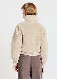 Giacca in pile beige, corta, con colletto alto e superficie testurizzata. Polsini e orlo a coste; indossata sopra pantaloni di velluto a coste marroni.