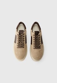 Baskets en suédine beige avec des accents en cuir marron, bout rond et lacets marron. Semelle en caoutchouc texturée, design minimal.