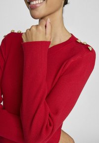 Sarkans ribots džemperis ar garajām piedurknēm, kuram ir zelta dekoratīvas pogas uz pleca un V-veida kakla izgriezums. Gluda tekstūra un pieguļošs siluets.