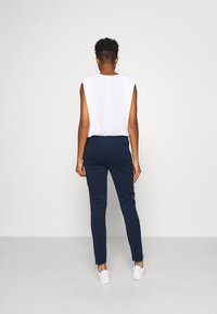 Vitt ärmlöst plagg, marinblå slim-fit byxor med vita sidostråk, bärs med vita skor. Slät tyg, avslappnad design, bakifrån.