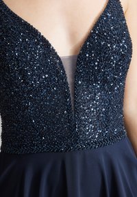 Sinivihreä mekko syvällä V-päiväkauluksella, jonka bodice on koristeltu paljeteilla, ja sujuva hame, joka laskeutuu vyötäröltä.