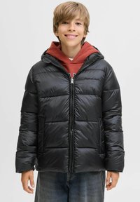 Doudoune noire avec capuche, design matelassé, fermeture éclair à l'avant et poches latérales, portée par-dessus un sweat à capuche rouge et assortie à un jean.